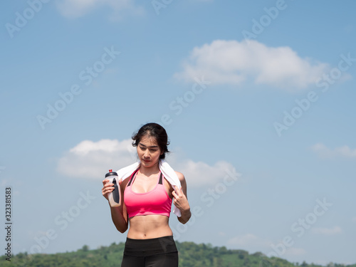 Front view Young asian woman running on road in the morning sunshine outdoors. Female jogger exercising in nature outdoors holding bottled water. Fitness and healthy lifestyle