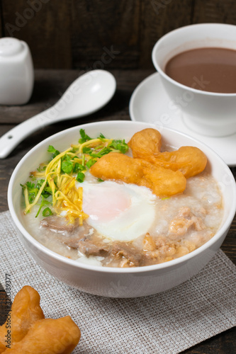 Rice gruel or Rice porridge with pork, onsen egg and deep fried dough stick in white bowl on wooden table.