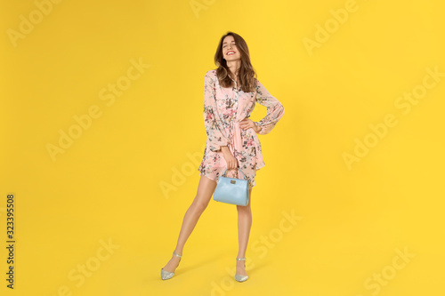 Fotografia Young woman wearing floral print dress with stylish handbag on yellow background