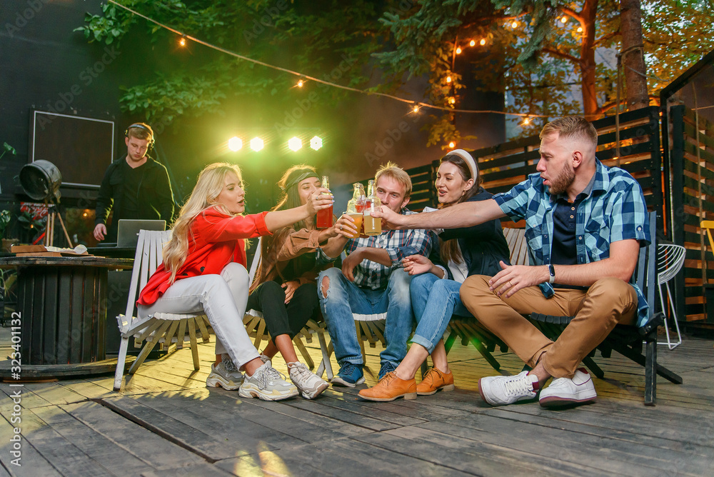 Fototapeta premium Friends clinking bottles and drink beer at outdoors cozy party with music and colored disco lights.
