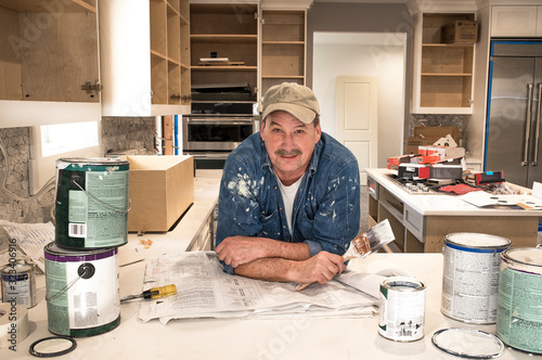 Male painter splattered with paint and holding a wet paint brush in messy home kitchen during residential home remodel with empty paint cans stacked around him, cabinet doors off and open