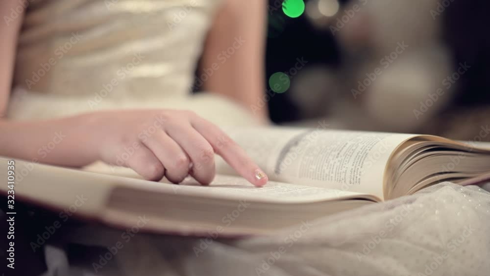 Close-up A little blonde girl in a festive dress with a book in her hands sits next to soft toys against the background of a Christmas tree and reads a book leading the page with her place.