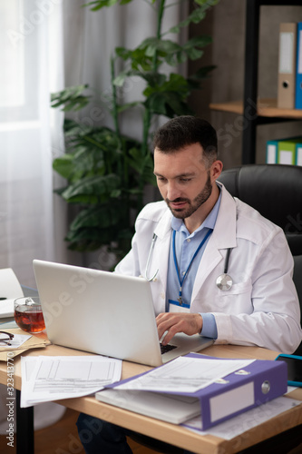 Young bearded doctor in a white robe working on laptop