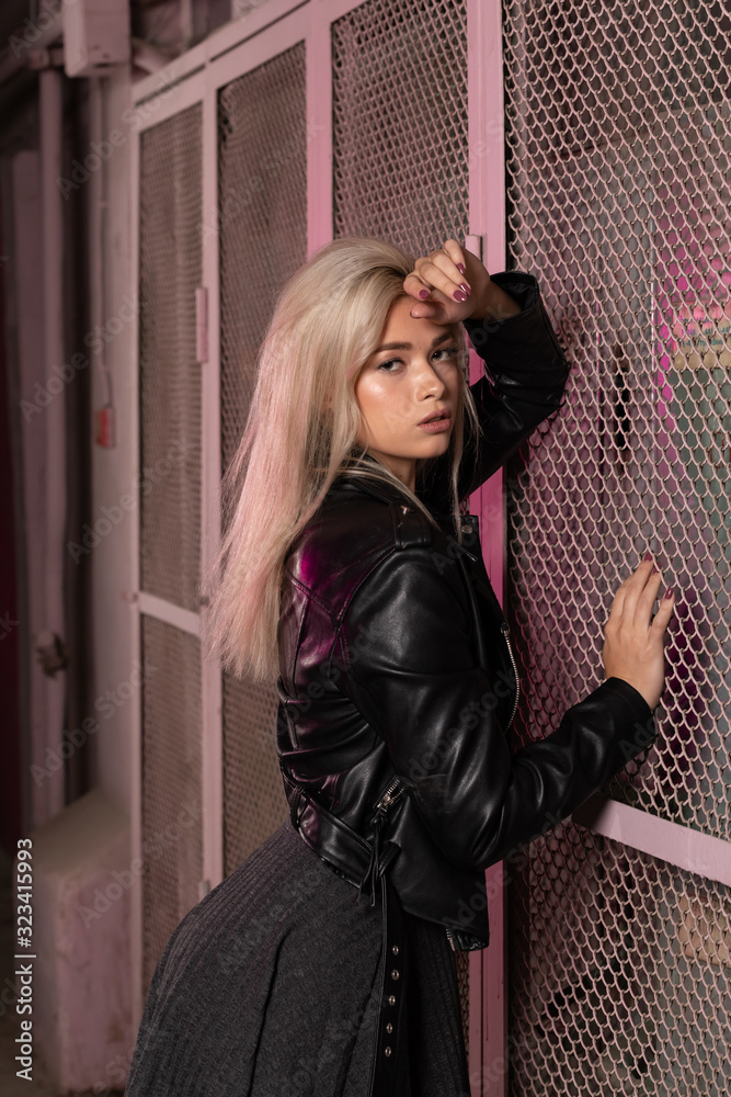 blonde girl in a black leather jacket posing on a dark background and pink lattice, rock style
