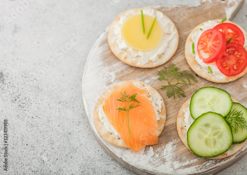 Various round healthy crackers with salmon and cheese, tomato and cucumber on wooden chopping board on light kitchen table background. Space for text