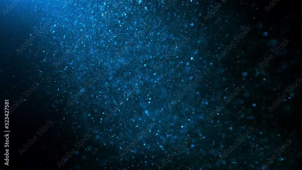 Blue glitter background in slow motion. Underwater particles flying and shining in bright spot light, shot with depth of field. Space dust beautiful bokeh abstract background