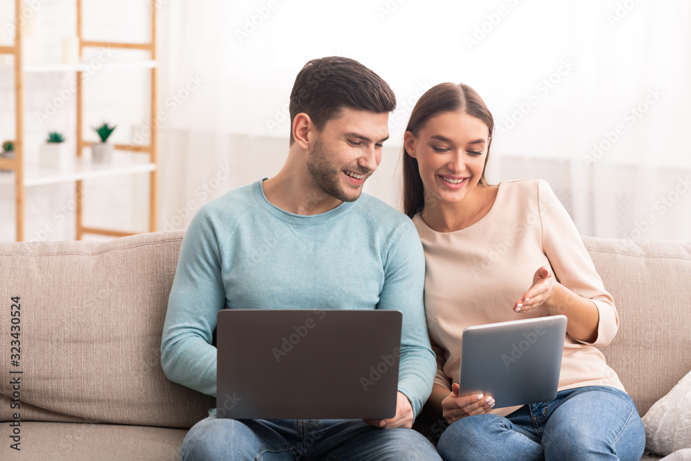 Couple Using Laptop And Tablet Sitting On Sofa At Home
