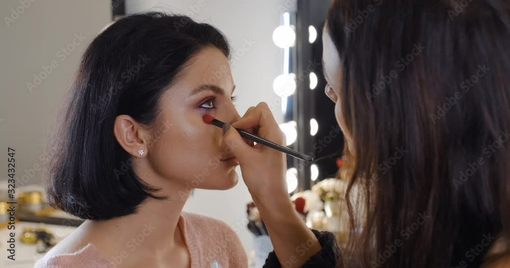 Portrait of brunette girl in makeup studio, makeup artist applies eye shadow with brush dipping into palette against background of bokeh light bulbs on mirror slow motion. Beauty lamp. Makeup Portrait of brunette girl in makeup studio, makeup artist applies eye shadow with brush dipping into palette against background of bokeh light bulbs on mirror slow motion. Beauty lamp. Makeup