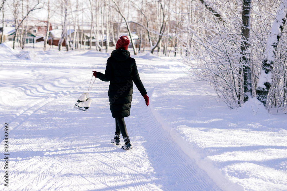 A young brunette in warm clothes with skates in her hands, against the backdrop of a city park full of snow. Winter vacation concept.