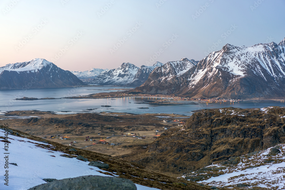 Obraz premium Landscape shot from Ryten towards fredvang and ramberg in Lofoten island in Norway during blue hour. Snow cover the peaks and city lights in the background.
