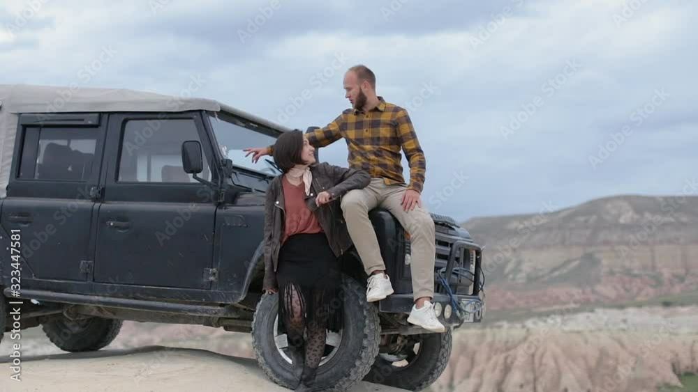 Young White Couple On A Road Trip By Car. Man Sitting On Hood Of Jeep Young White Couple On A Road Trip By Car. Man Sitting On Hood Of Jeep