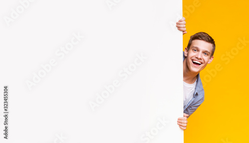 Emotional young guy peeking out from advertising empty board photo