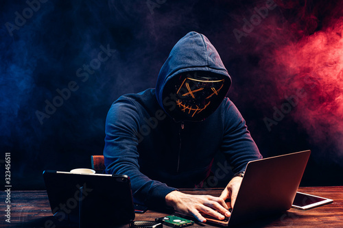 young criminal male hides his face under the hood and mask, hacks the password on the laptop, typing something. anonymous, incognito guy going to hack. cyberattack concept
