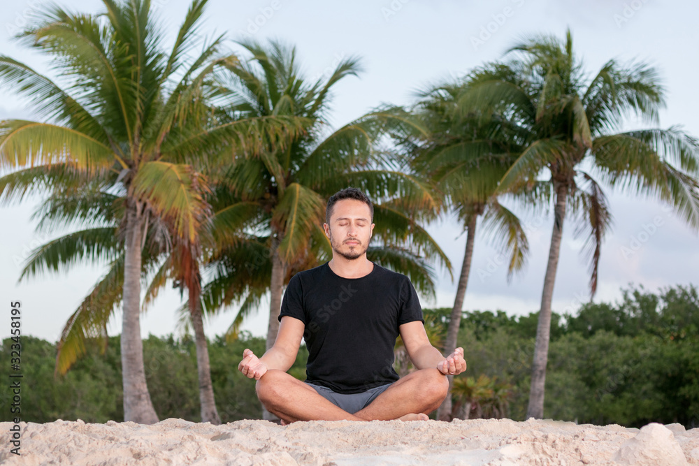 Young yoga man wearing black t-shirt at the beach, relaxing, breathing and meditating in a natural environment with palm trees and ocean as background.  - obrazy, fototapety, plakaty 