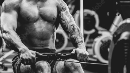 close up Young muscular man with tattoo doing exercises on rowing machine in gym, dramatic black and white image