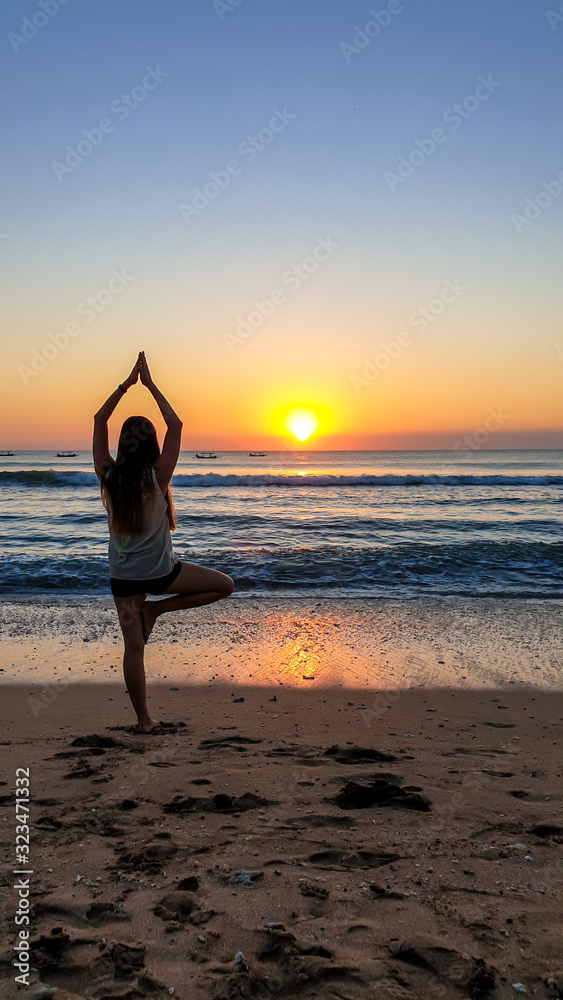 A girl practising yoga during the sunset on Seminyak beach on Bali, Indonesia. The sun sets directly into the water. Calm sea washes gently the shore. The sunbeams reflecting on the sea surface.