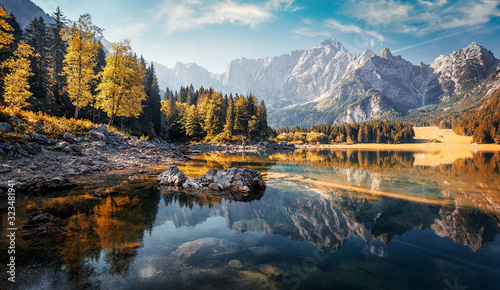 Awesome sunny landscape in the forest. Wonderful Autumn scenery. Picturesque view of nature wild lake. Sun rays through colorful trees. Incredible view on Fusine lakeside. Amazing natural Background