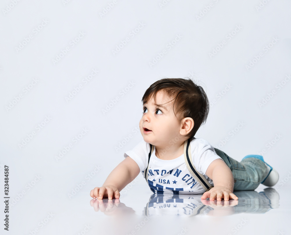 Little toddler in t-shirt with inscriptions, jeans with suspenders. He is laying on floor isolated on white background. Close up Little toddler in t-shirt with inscriptions, jeans with suspenders. He is laying on floor isolated on white background. Close up