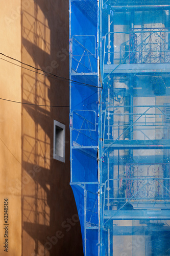 Immeuble en construction avec échafaudage protégé par un filet de sécurité bleu projetant son ombre sur un immeuble voisin ocre jaune.