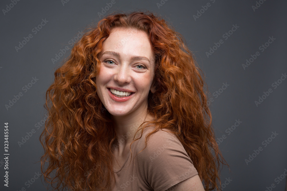 Happy curly red-haired woman with beautiful smile, holding her hair with her hands. On an isolated gray background. Concept: happiness and emotions Happy curly red-haired woman with beautiful smile, holding her hair with her hands. On an isolated gray background. Concept: happiness and emotions