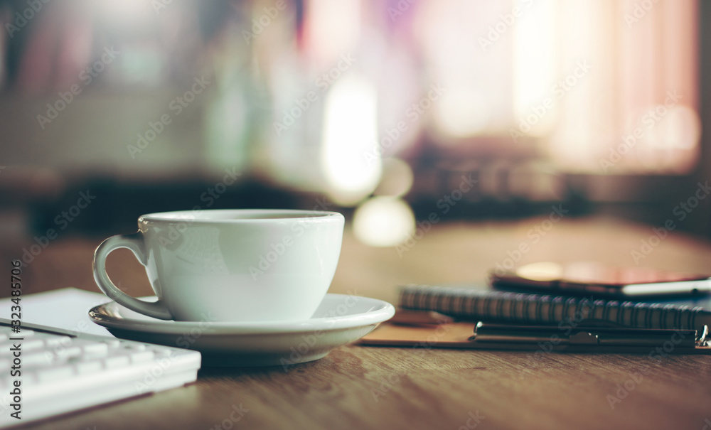 Closeup white cup of coffee with smartphone, notebook  on wooden table in cafe. with vintage light, blurred and bokeh background