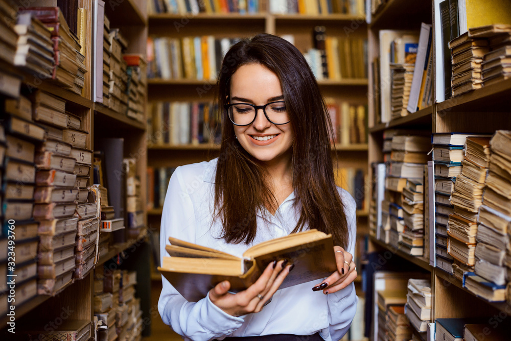 Cheerful brunette female student reading book in the library, standing between aisles of shelves of different old books Cheerful brunette female student reading book in the library, standing between aisles of shelves of different old books