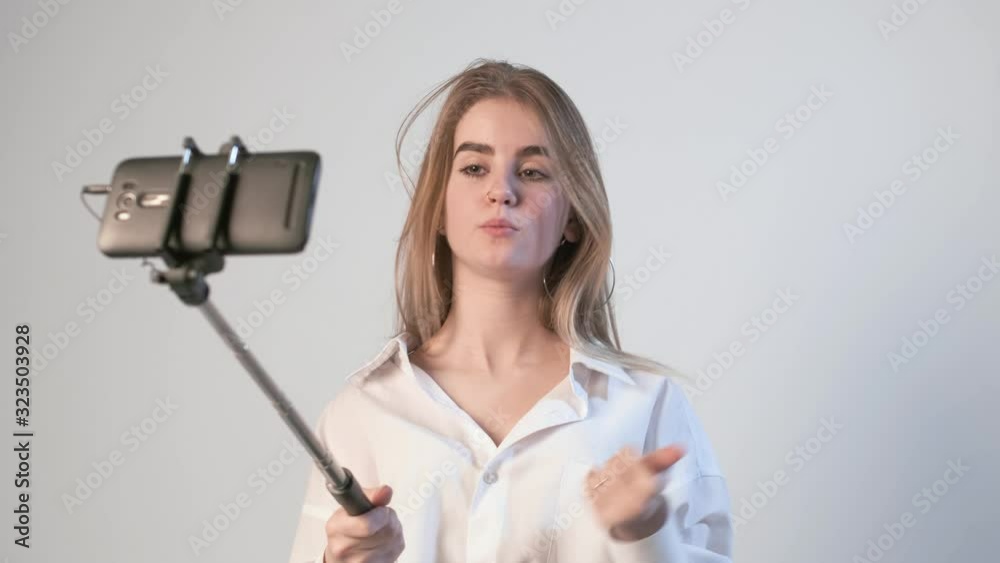 young female records video with smartphone attached to selfie stick. blond girl filming tutorial vlog about eyebrows cosmetics and makeup, using cellphone isolated on white background. Beauty blogger young female records video with smartphone attached to selfie stick. blond girl filming tutorial vlog about eyebrows cosmetics and makeup, using cellphone isolated on white background. Beauty blogger