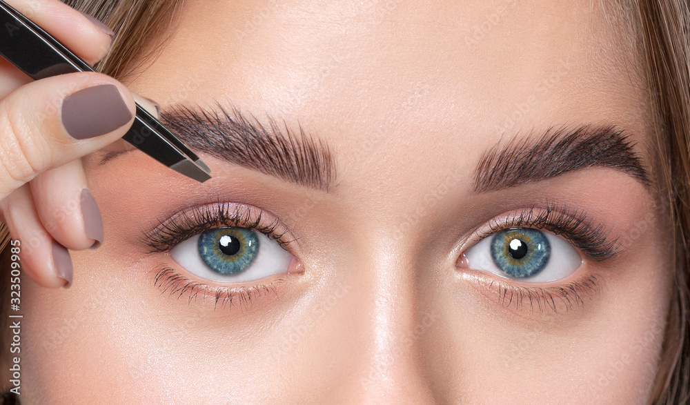 Make-up artist plucks eyebrows with tweezers to a woman with curly brown hair and nude make-up. Beautiful thick eyebrows close up. Professional makeup and cosmetology skin care.