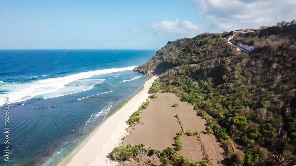 A done shot of Nyang Nyang Beach, Bali, Indonesia. The waves are rushing to the shore, making the water bubbly. The beach is covered with green algae, further on it's sandy. Tall cliffs on the side