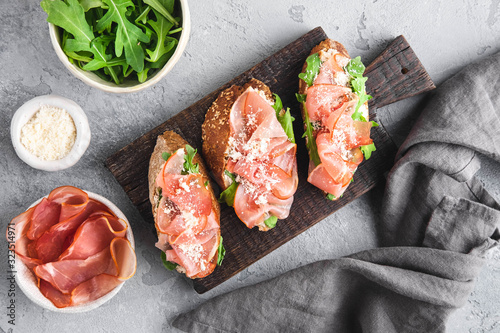 Italian appetizer bruschetta with parma ham, arugula and grated parmezan on a wooden board on a grey background, close up top view, horizontal