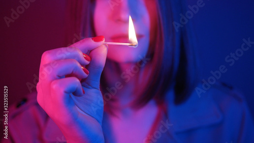 Attractive young woman holding matchstick with light in fingers, blowing out, looking at camera. Blue and purple combination of lights. Bob hairstyle. Red nails.