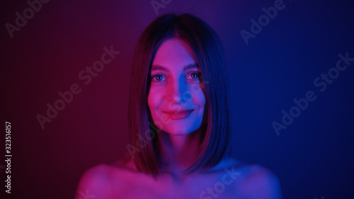 Shy gorgeous young woman with straight hair looking into camera, smiling. Footage of lovely Caucasian girl with beautiful collarbone in blue and purple lights. Bob hairstyle.