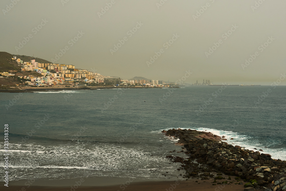 Day with calm, where you can see in the foreground part of the beach with breakwaters and calm sea. In medium plane, buildings of different heights and overpasses. In the background you can see part o
