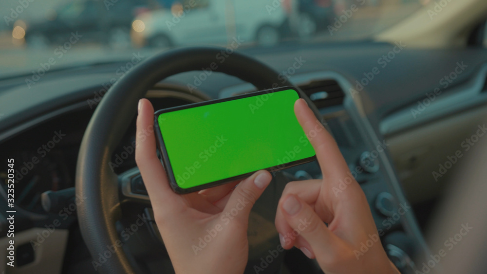 Lviv, Ukraine - May 19, 2018: Female driver sitting in the car browsing online map on vertical mock-up smartphone greenscreen searching route planning a roadtrip. Lifestyle and technology.