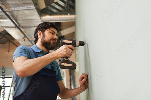Low angle portrait of bearded construction worker drilling wall while renovating house alone, copy space photo