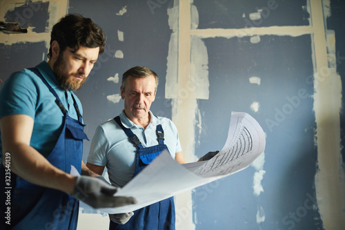 Portrait of two construction workers holding plans and standing against dry wall while renovating house, copy space photo