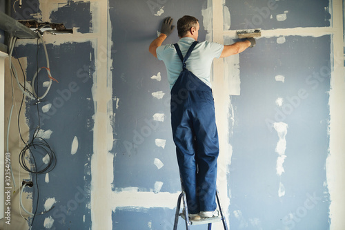 Full length back view of senior construction worker smoothing dry wall standing on ladder while renovating house, copy space photo