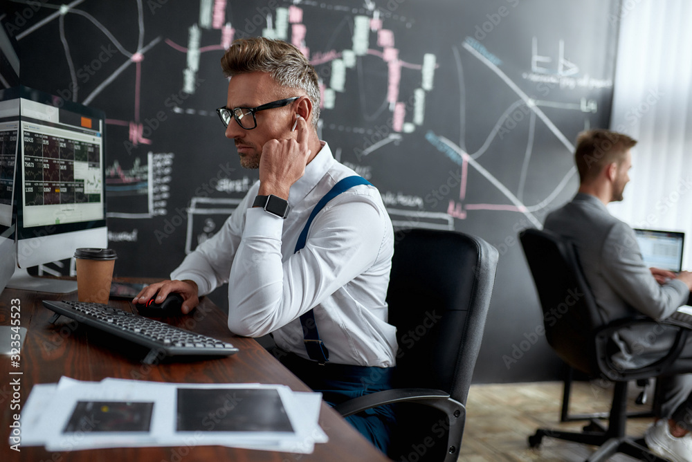 Satisfying the needs. Stylish businessman, trader sitting in front of computer monitor while working using wireless earphones. His colleague and blackboard full of data analyses in the background. Satisfying the needs. Stylish businessman, trader sitting in front of computer monitor while working using wireless earphones. His colleague and blackboard full of data analyses in the background.