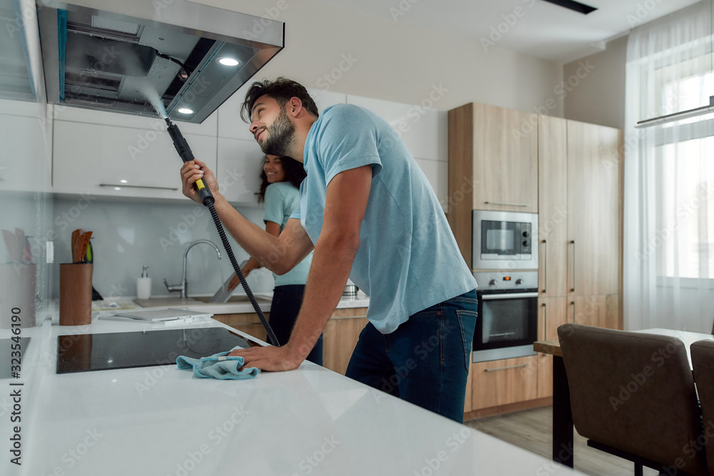 © Svitlana - Young caucasian man in uniform cleaning kitchen range hood with steam cleaner while his smiling female colleague washing something in the sink on the background. Cleaning services concept © Svitlana - Young caucasian man in uniform cleaning kitchen range hood with steam cleaner while his smiling female colleague washing something in the sink on the background. Cleaning services concept