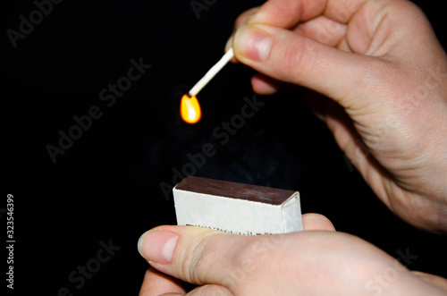 A hand holds a burning match on a black background. A wooden match burns in the hands of a macro. Igniting a match on a box. Smoke from a match.  Flash