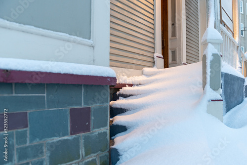 The steps of a vintage door filled with drifts of snow. The building is tan in color with white trim. The basement wall is made of green and purple slate rock. The drift covers all the steps upward.