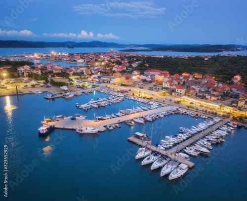 Aerial view of boats and yachts in port and city at night. Summer landscape with city lights, buildings, illuminated streets, mountain, motorboats, blue sea, sky at dusk. Top view. Travel in Croatia
