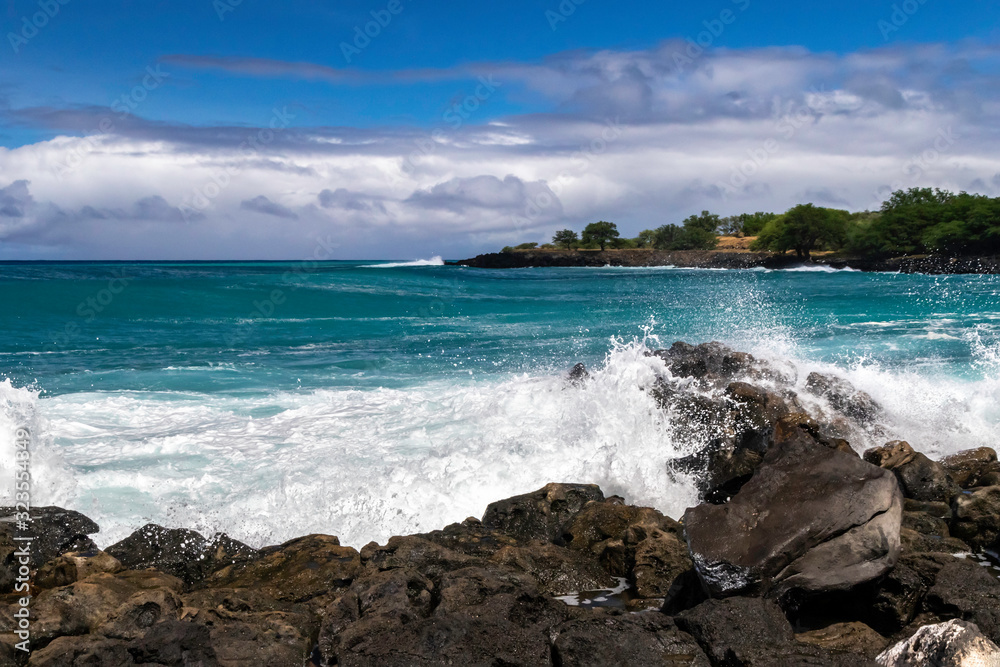 Wave breaking on rough shore of the Kona coast, on Hawaii's Big Island. Blue-green Pacific ocean beyond; rocky shoreline with trees in the distance. Cloudy blue sky above.