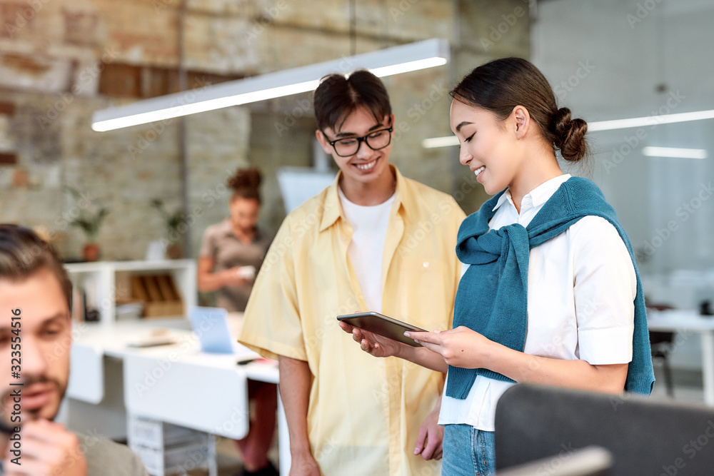 We did it. Two asian colleagues in casual wear looking at digital tablet and discussing something while standing in the creative office We did it. Two asian colleagues in casual wear looking at digital tablet and discussing something while standing in the creative office