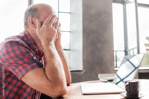 Tired caucasian senior bald man rubbing his eyes near laptop computer sitting in the living room.