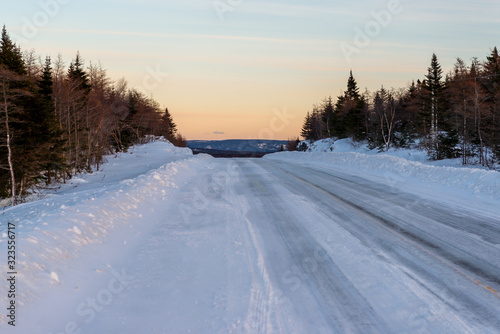A snow covered country road with the sun setting in the distance. Trees line the sides of the road. There's a worn tire pattern in the snow. The sun has just started to set and is a pale orange color.