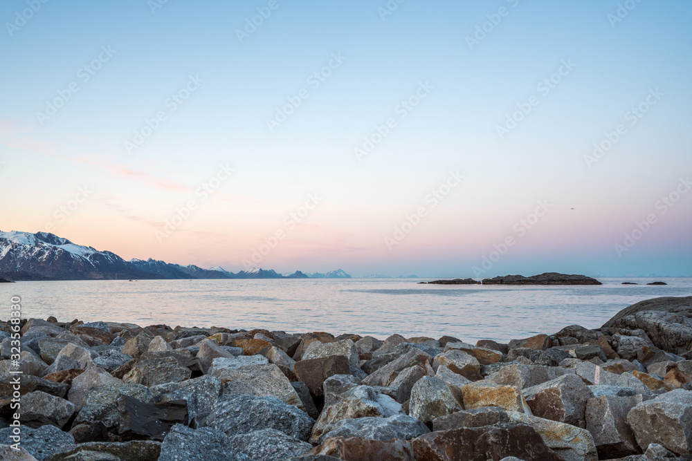 Overlooking the ocean outside Hamnøy in Lofoten, Norway during blue hour. Pink and blue tones over snow covered mountains. Wave breaker boulders as foreground.