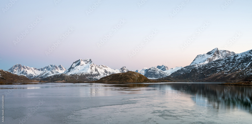 Panorama of Snow covered mountains and powerful streams in the ocean during blue hour in Flakstad, Lofoten in Norway. Pink and blue tones in the sky.