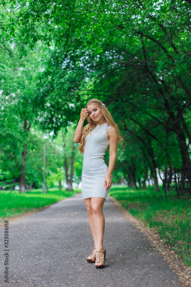 Portrait of a charming blond woman wearing beautiful white dress standing on the road under trees. Portrait of a charming blond woman wearing beautiful white dress standing on the road under trees.