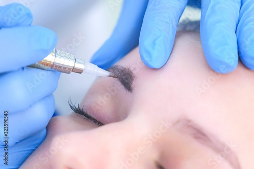 Cosmetologist making eyebrows microblading procedure in beauty salon for girl using tattoo apparatus, eyebrow closeup. Beautician in gloves is doing permanent makeup to woman. Beauty industry concept.
